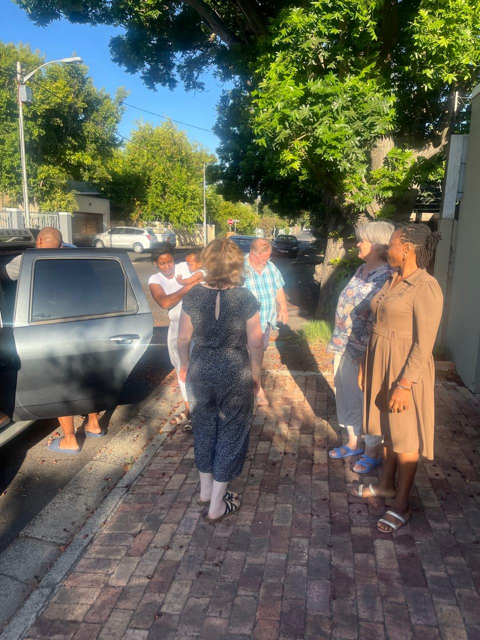 Brothers and sisters greeting each other on a tree-lined Cape Town street as John arrives by car