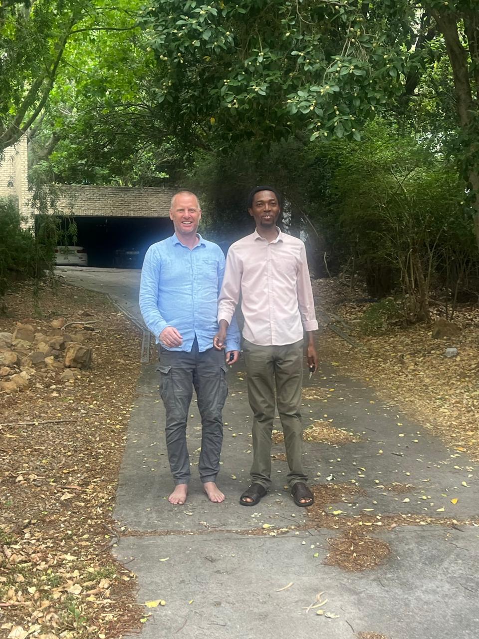John Damilola standing with a brother on a driveway beneath large shady trees